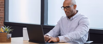 A person wearing glasses is seated at a table, using a laptop