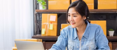 A woman in a denim shirt uses a laptop at a desk cluttered with cardboard boxes.