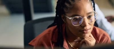  Person with braided hair wearing glasses and a rust‑colored shirt, seated at a desk.