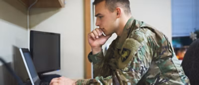 A person in military uniform focused on a laptop screen in an office setting.