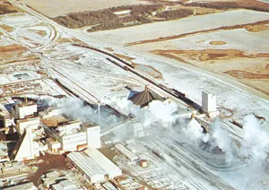 Aerial view of the potash mine at Esterhazy, Saskatchewan, showing industrial buildings, storage facilities, and plumes of steam or smoke surrounded by snowy fields.