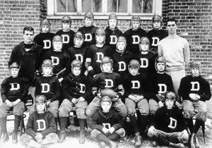 Sepia-toned group photo of a high school football team with boys in vintage uniforms and leather helmets, including John F. Kennedy, posing in front of a brick building.