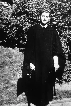 John F. Kennedy standing outdoors wearing dark university graduation robes, with greenery in the background.