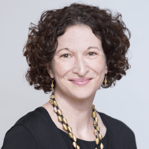 Woman with curly brown hair wearing a black top, gold earrings, and a necklace, smiling against a light gray background.