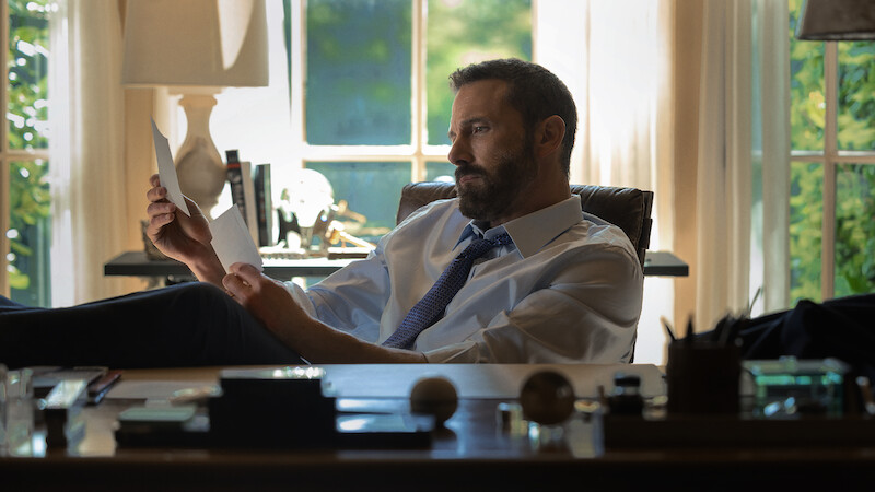 A man in a shirt and tie sits at a desk in a sunlit office, reading a document with his feet up. The office has large windows, books, and various desk items visible in the background.