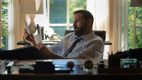 A man in a shirt and tie sits at a desk in a sunlit office, reading a document with his feet up. The office has large windows, books, and various desk items visible in the background.