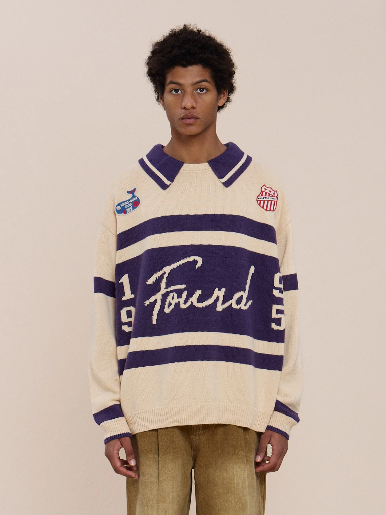 Someone wears the FOUND Knit Jersey with cream and purple stripes, “Found” and “1995” on the front, embroidered patches, and tan pants against a plain beige background.