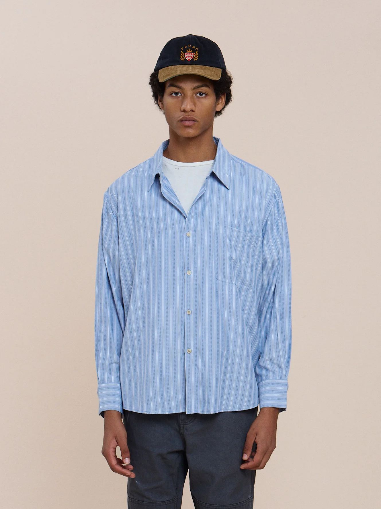 Model wears the FOUND Stripe Pocket Oxford Shirt, unisex relaxed fit, layered over a white tee and dark pants against a light background.
