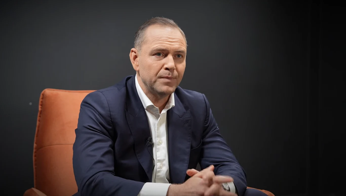 Karol Nawrocki odniósł się do sprawy mieszkania Jerzego Ż. Mężczyzna w garniturze siedzi na pomarańczowym fotelu, patrzy w stronę aparatu, złożył ręce w geście spokoju. Ciemne, neutralne tło nie odwraca uwagi od postaci.