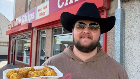A picture of Kalani holding a tray of 'spicy spuds'. He is wearing glasses and a black hat.