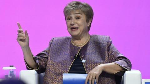 Kristalina Georgieva, managing director of the International Monetary Fund, sits in an armchair, speaking and smiling while pointing upwards. The background is pink, and she wears a shiny jacket with a gold diamond repeated motif.