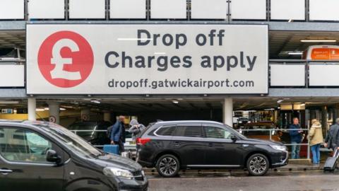 A drop-off zone at Gatwick Airport, showing two cars parked up and passengers walking to terminals. There is a huge sign saying charges apply.