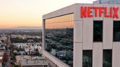 A wide view shows the Netflix logo on the side of a cream coloured building at Netflix studios in 2025 in LA, with the iconic LA skyline in the background at sunset.