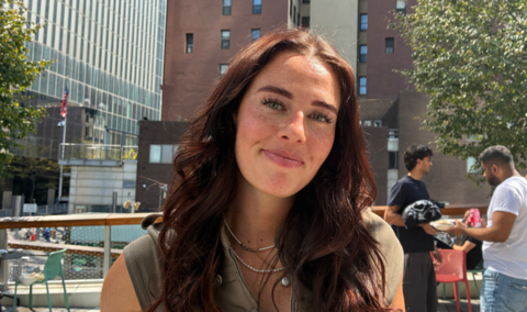 Willa, a young woman with long brown hair standing outside in front of buildings, smiling at the camera