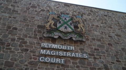 Outside of Plymouth Magistrates' Court. It is a close up of the brick wall with a large sign with the words 'Plymouth Magistrates Court' with a large badge above it.