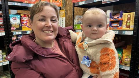 A woman with brown hair tied back in a ponytail is wearing a dark pink padded jacket. She is standing in front of shelves holding a baby with a bald head and wearing a cream fleece with orange pumpkins and a blue dummy