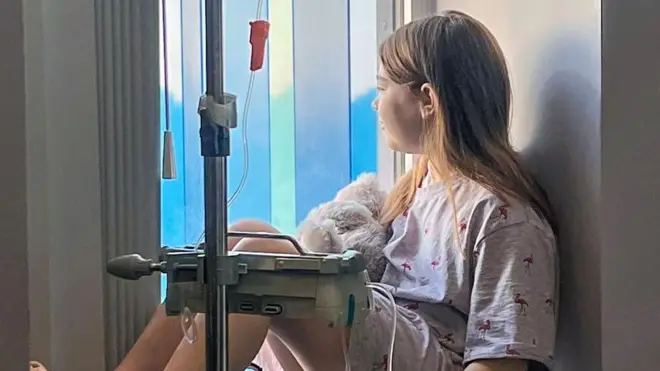 A little girl of around 13 years old with long straight brown hair tucked behind her ears sits half silhouetted in a hospital window attached to a drip with a teddy bear in her lap. She is looking away from the camera and out of the window.