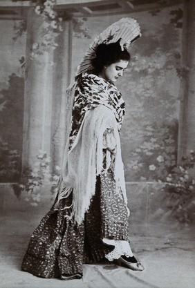 A standing woman, posed with an upraised fan and pointed toe, wearing a fringed shawl, in front of a painted backdrop in a photographic studio. Photograph, ca.1900.