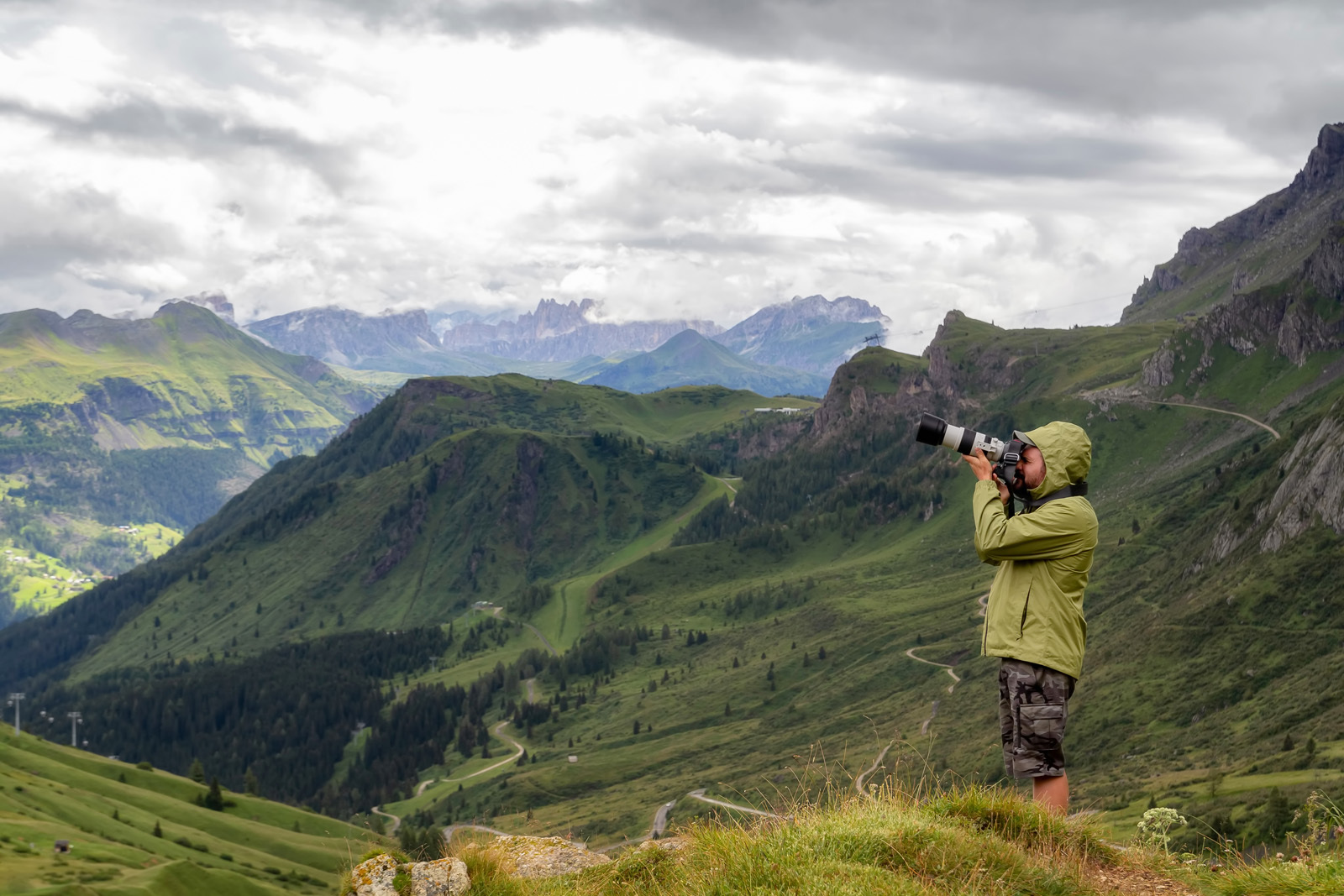 A side profile of a person wearing a coat and shorts, looking through a telephoto lens to photograph a mountainous landscape. Above is a slightly grey, cloudy sky.