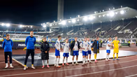 Ett fotbollslag i vita och blå matchställ står uppradade på en fotbollsplan framför tomma läktare.