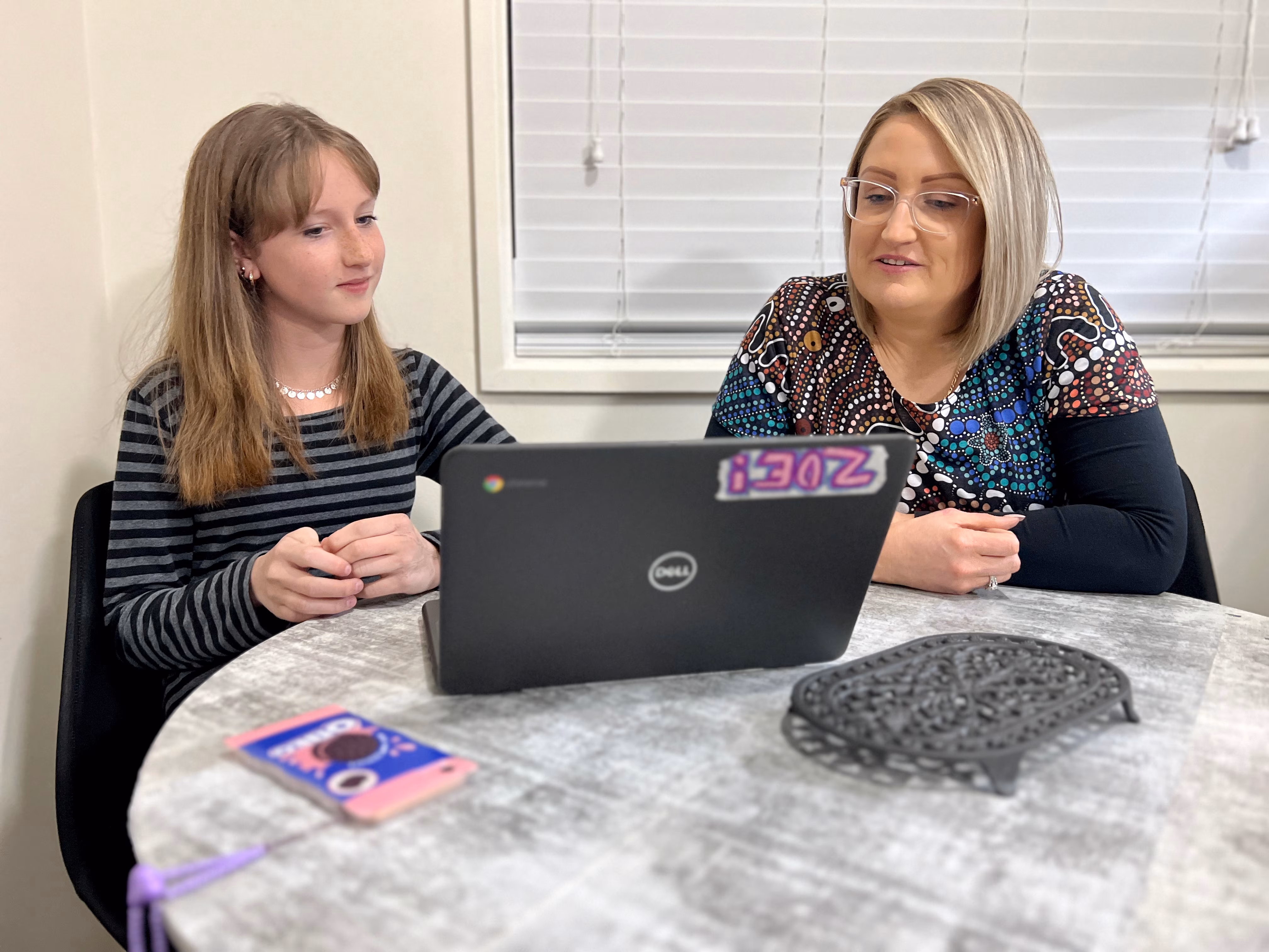 A girl and a woman sit at a round table indoors, looking at a laptop screen.
