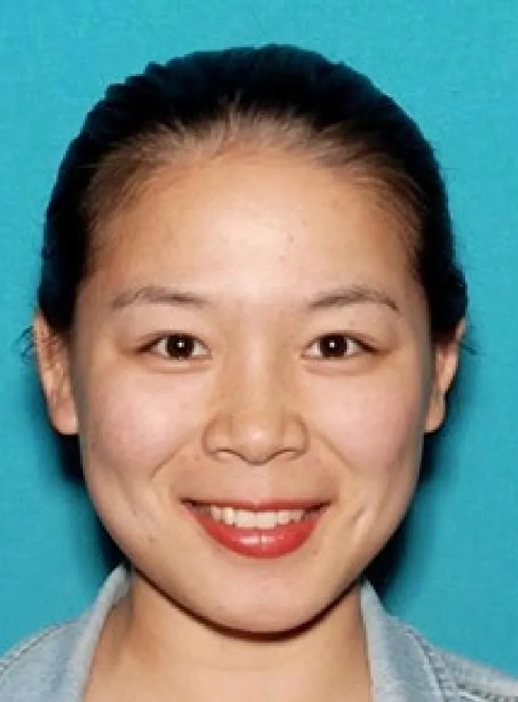 ID-style portrait of a young woman with dark hair pulled back, smiling against a blue background.