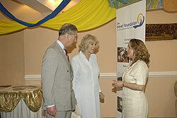 Charles wearing a suit converses with a woman in a dress. Camilla stands next to Charles in the background standing opposite a "Travel Foundation" vertical banner.