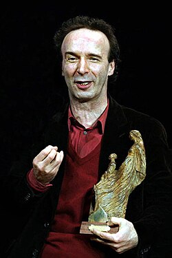 Portrait of a man holding a gold statuette. He is wearing a black coat over a red sweater and a red collared shirt.
