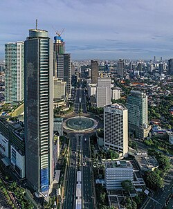 Aerial view of modern skyscrapers of Jakarta and a wide-city boulevard surrounding a large circular fountain roundabout amid a dense urban landscape under a clear blue sky.