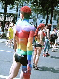 A muscular, young, man walking in a Pride parade in his underwear only and rainbow paint decorating his body.