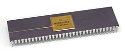 Close up of a small dark grey rectangular microprocessor chips with small gold-colored metallic prongs extending from the side downward. The top has a small gold plate in the middle that reads "MC68010L8, 1TA28552".