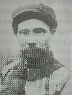 Man with a black cylindrical hat, thick black beard, rimless glasses, wearing a black tunic, stands in front of a white background.