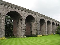 A series of seven stone archways, connected to stone parapet above grass area.