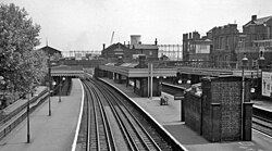 Black and white photograph of a four track station with an island and two side platforms. Behind the station building that is facing us in the background are a power station cooling tower and gasholders.