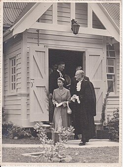Elizabeth II and a man in ceremonial robes stand outside a wooden building, with two men visible in the doorway behind them.