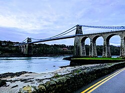 Pale blue metal suspension bridge carrying road traffic across a body of water with large stone arches at one end
