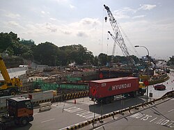 Construction site of Haw Par Villa station along a busy road, featuring large lattice boom cranes over an excavation pit surrounded by safety netting, steel sheet piles, and perimeter hoarding.