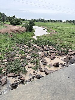 A high-angle view looking down at a rocky stream bed filled with large scattered boulders and turbid water. The left bank shows an eroded mound of reddish-brown earth.