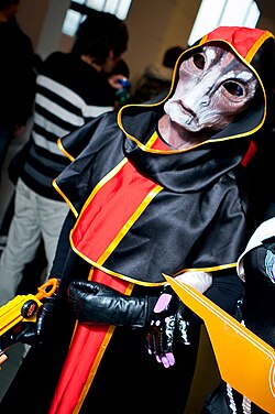 A person wearing a full-head mask meant to resemble the appearance of the species, similar to the one described in the infobox but, in reddish-purple. They are wearing a black and red cloak with a hood that covers the mask's horns.