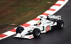 A figure, wearing a helmet of a dark blue design with red and yellow stripes, is driving a Formula One with an white colour scheme adorned with sponsorship. He is holding both hands on a steering wheel, turning right.