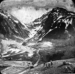 Black and white photo, a group of five people stand near a path that eventually winds between two snow-covered peaks.