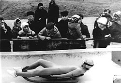 Photo en noir et blanc d'une lugeuse pendant une course, devant des spectateurs.