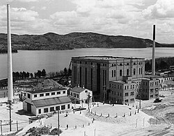 Large building shaped like a cube. In front is a smaller, barn-like building. They are flanked by two smokestacks. There is a river in the background.
