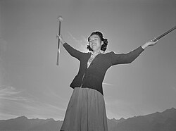Japanese teenage girl in 1940s sweater, skirt, and blouse twirling two batons and smiling, backlit by the sun against a nearly-cloudless sky.