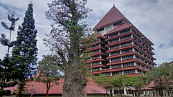 The University of Indonesia rectorate, a tall tiered building with a steep pyramidal roof, framed by trees and lower red-roofed buildings.
