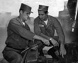 "Two men in military uniform at an Army base, ponder over instructions on how to change parts of a military truck."