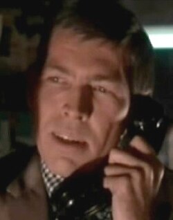Headshot of a man holding a black telephone. He is wearing a checkered shirt and a dark brown suit.