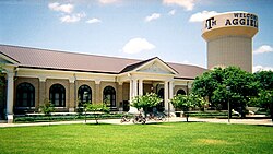 A neoclassical building with columns in front of a water tower sporting the greeting, "Welcome to Aggieland"