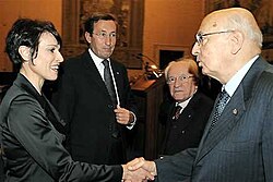 Photographie de quatre personnages en tenue de ville, une femme aux courts cheveux bruns serrant la main d'un homme dégarni portant des lunettes, deux autres hommes les regardant, en retrait.
