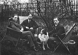 Les frères Duchamp : les trois hommes, en costume de ville, sont photographiés assis dans des fauteuils ; un chien border collie est assis à leurs pieds.